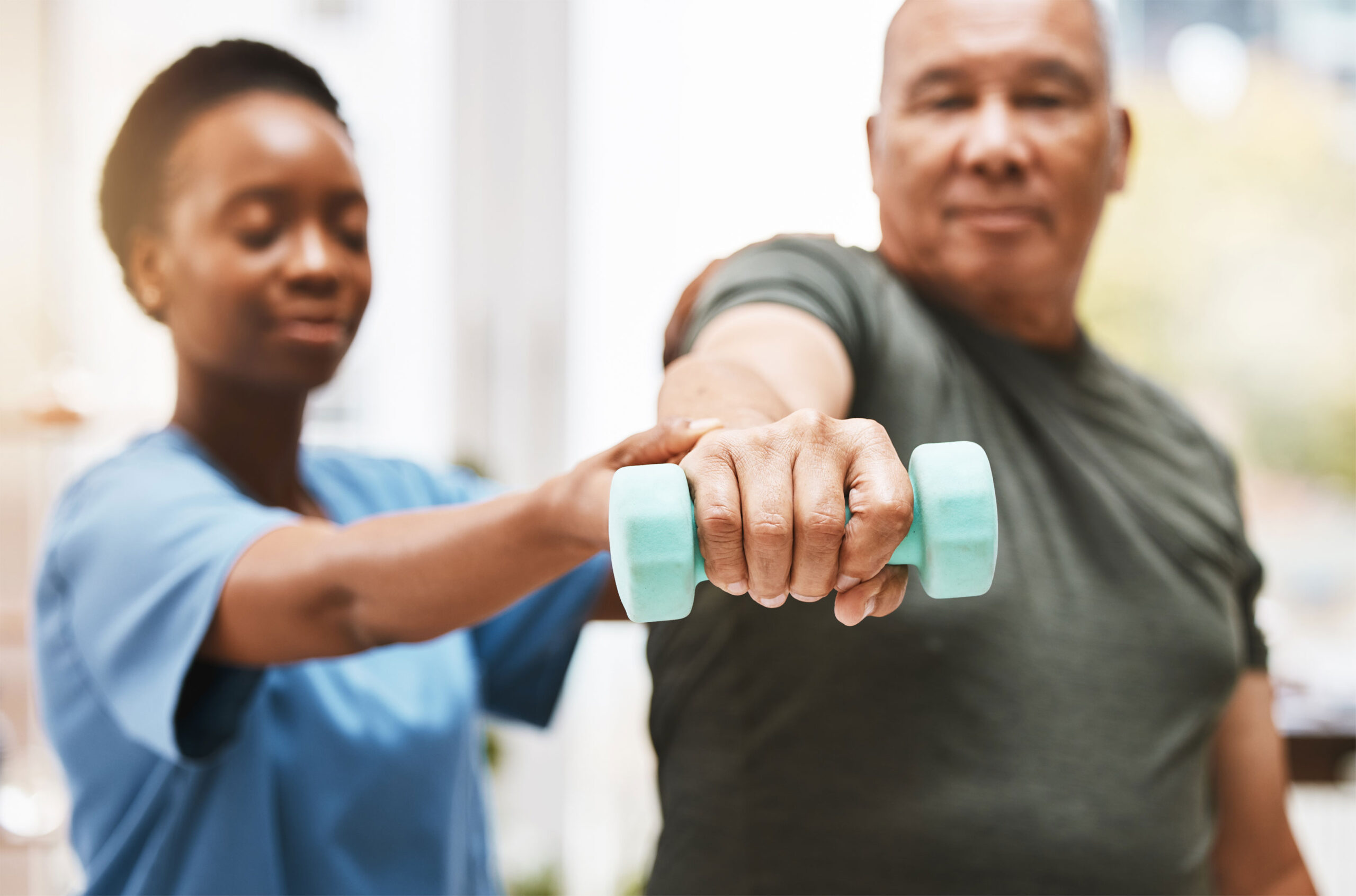 Middle age man holds a light dumbbell in his outstretched arm while therapist stands next to him and lightly supporting his wrist holding the weight.