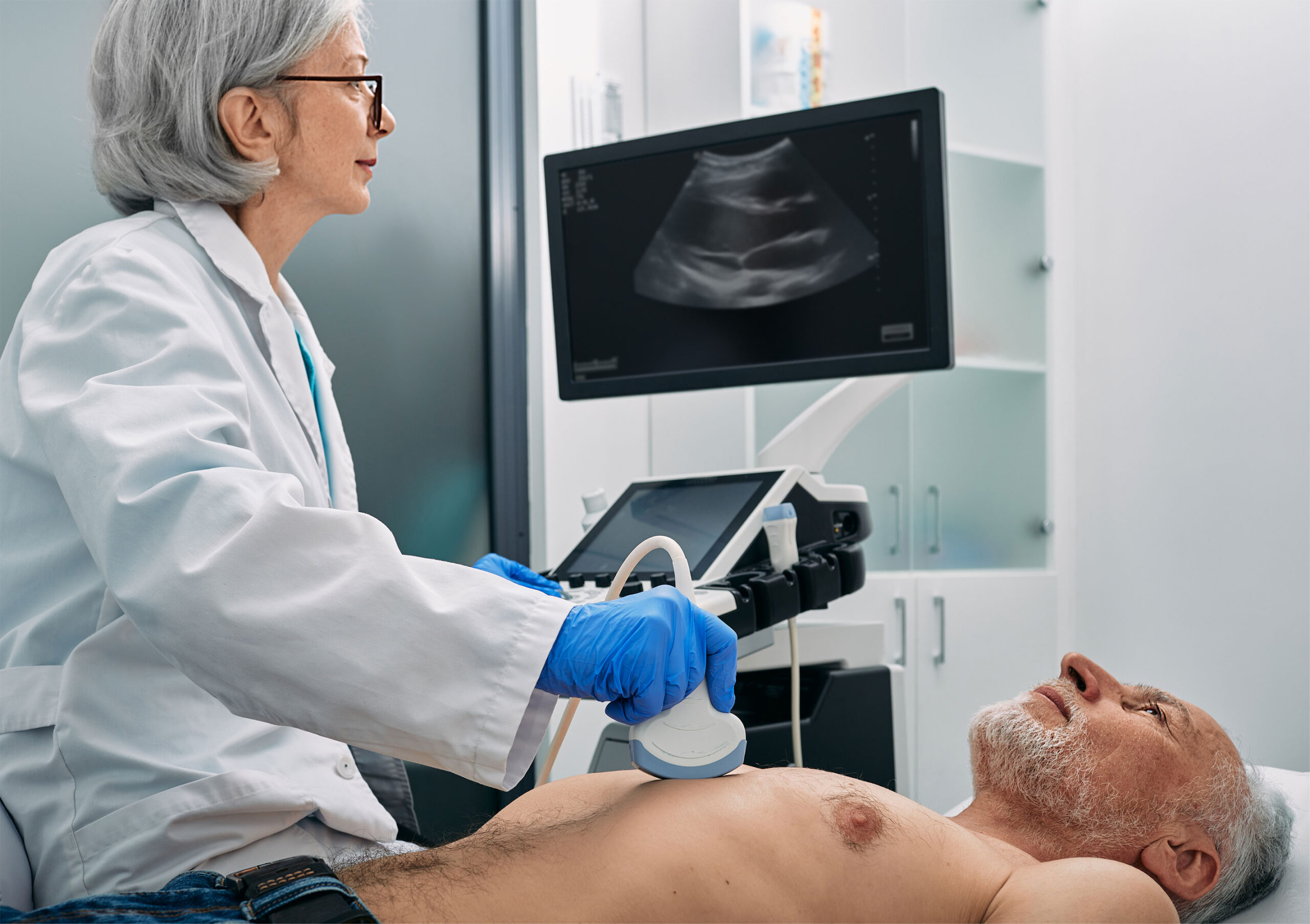 Senior man on examining table while technician holds an ultrasound wand over his heart with diagnostic machine in the background.